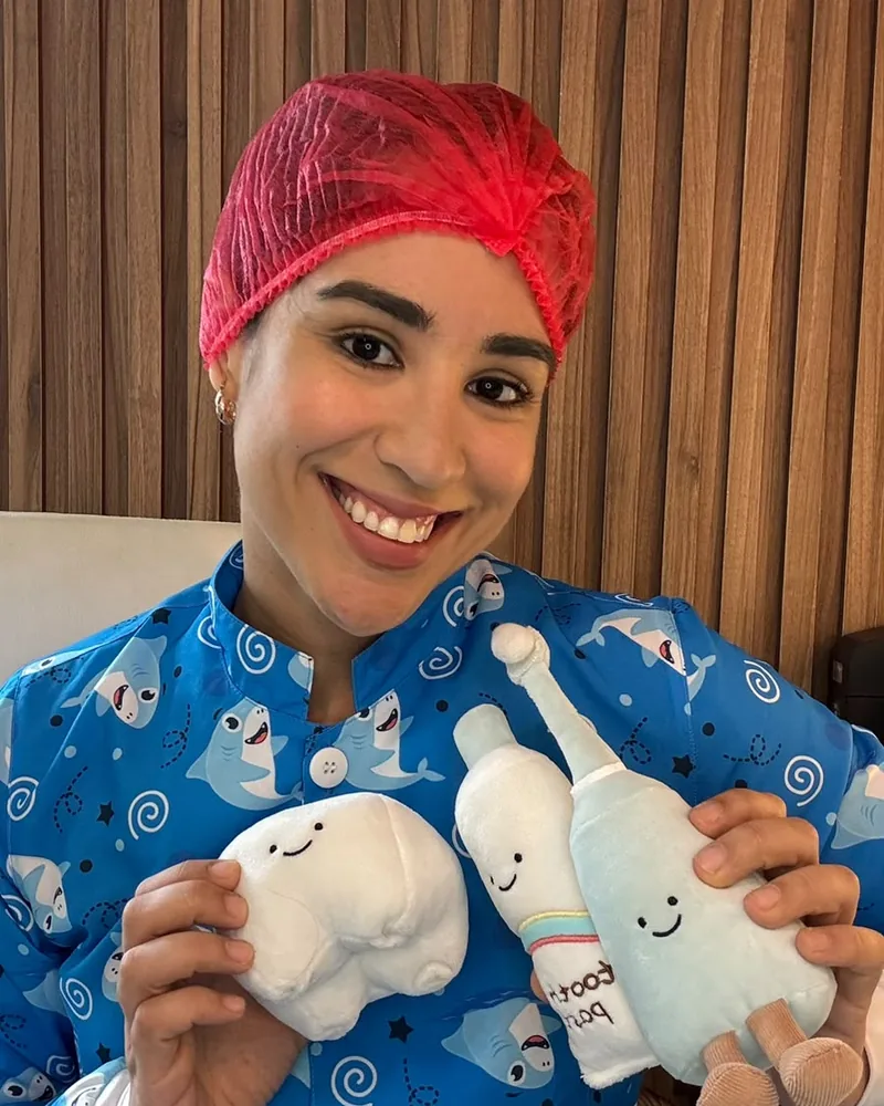 Dra. Milena Amancio sorrindo, segurando pelúcias de dentinho, escova e creme dental